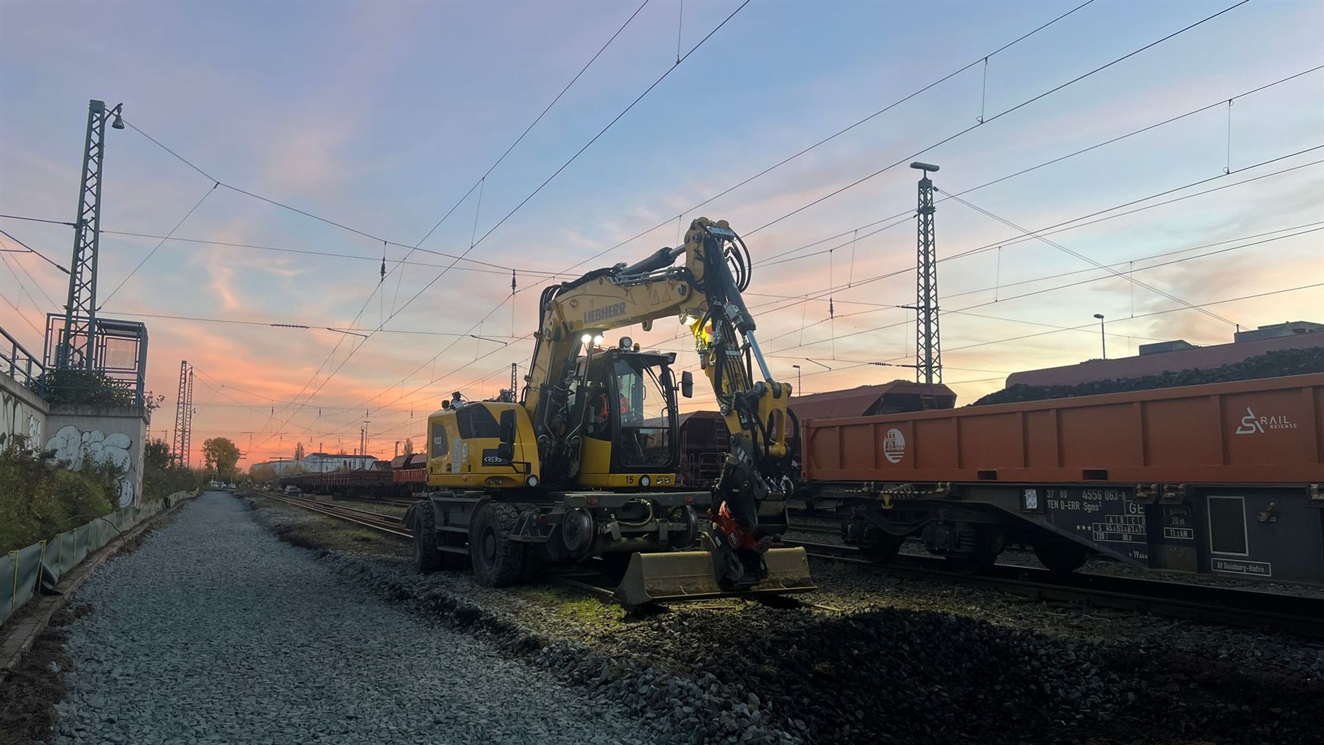 Bagger neben Güterwagen und Gleisen in der Abenddämmerung.