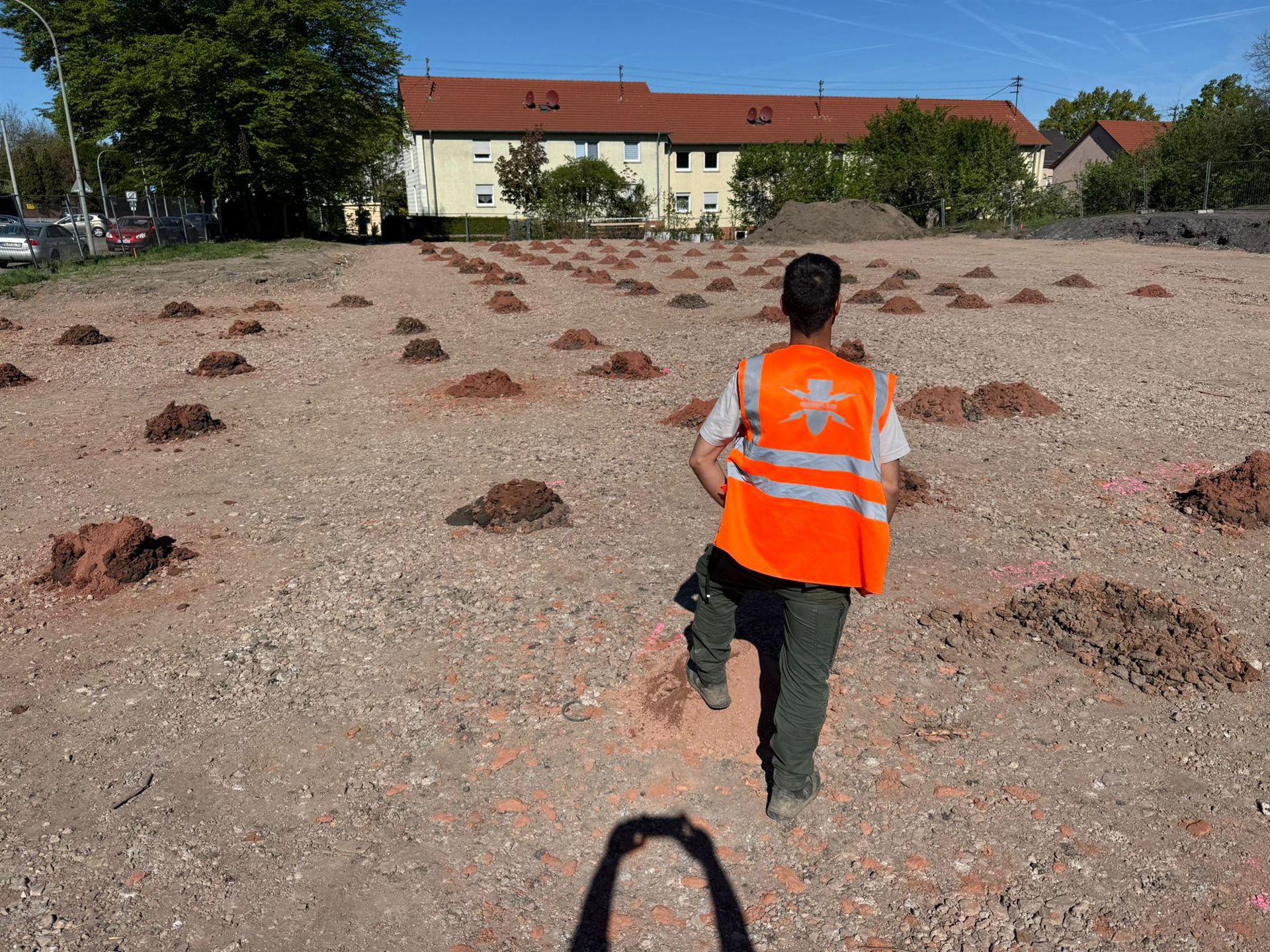 Person in orangefarbener Warnweste auf einer markierten Baustellenfläche mit kleinen Erdhaufen