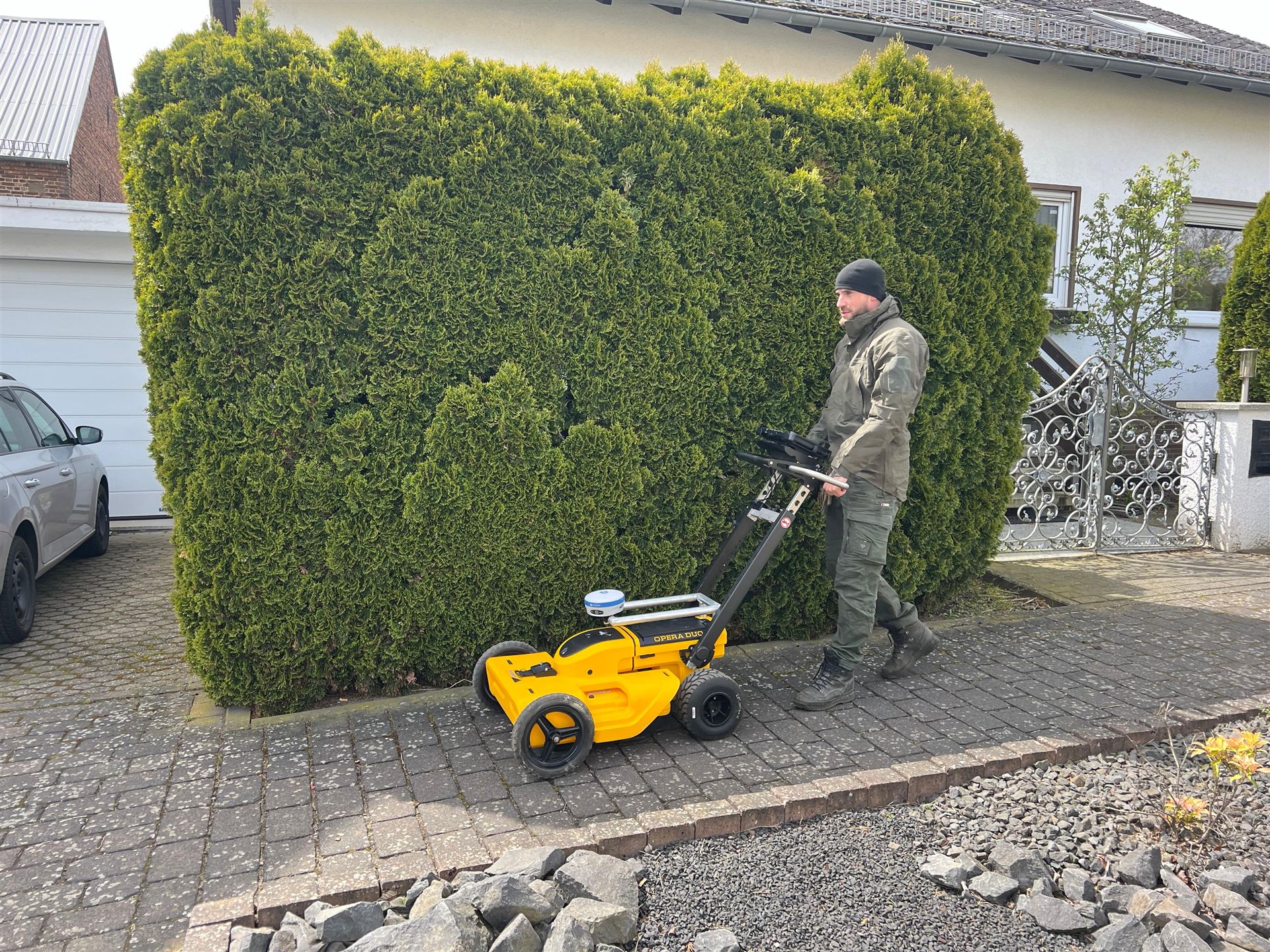 Person mit gelbem Messwagen auf einem gepflasterten Gehweg vor einer Hecke.