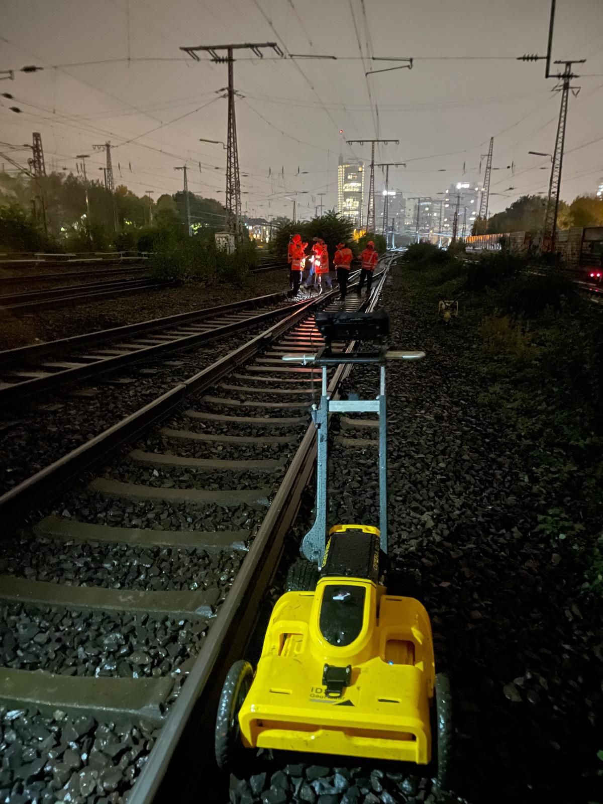 Gelber Messwagen auf einem Gleis bei Nacht, dahinter Personen in Warnkleidung.