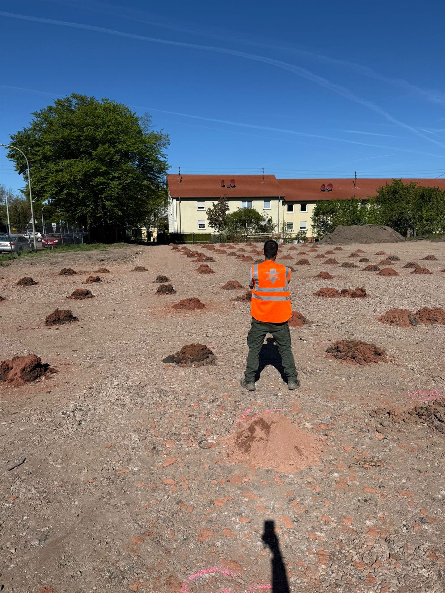 Person in orangefarbener Warnweste auf einer offenen Bodenfläche mit kleinen Erdhaufen und rosa Bodenmarkierungen; im Hintergrund stehen Wohngebäude.