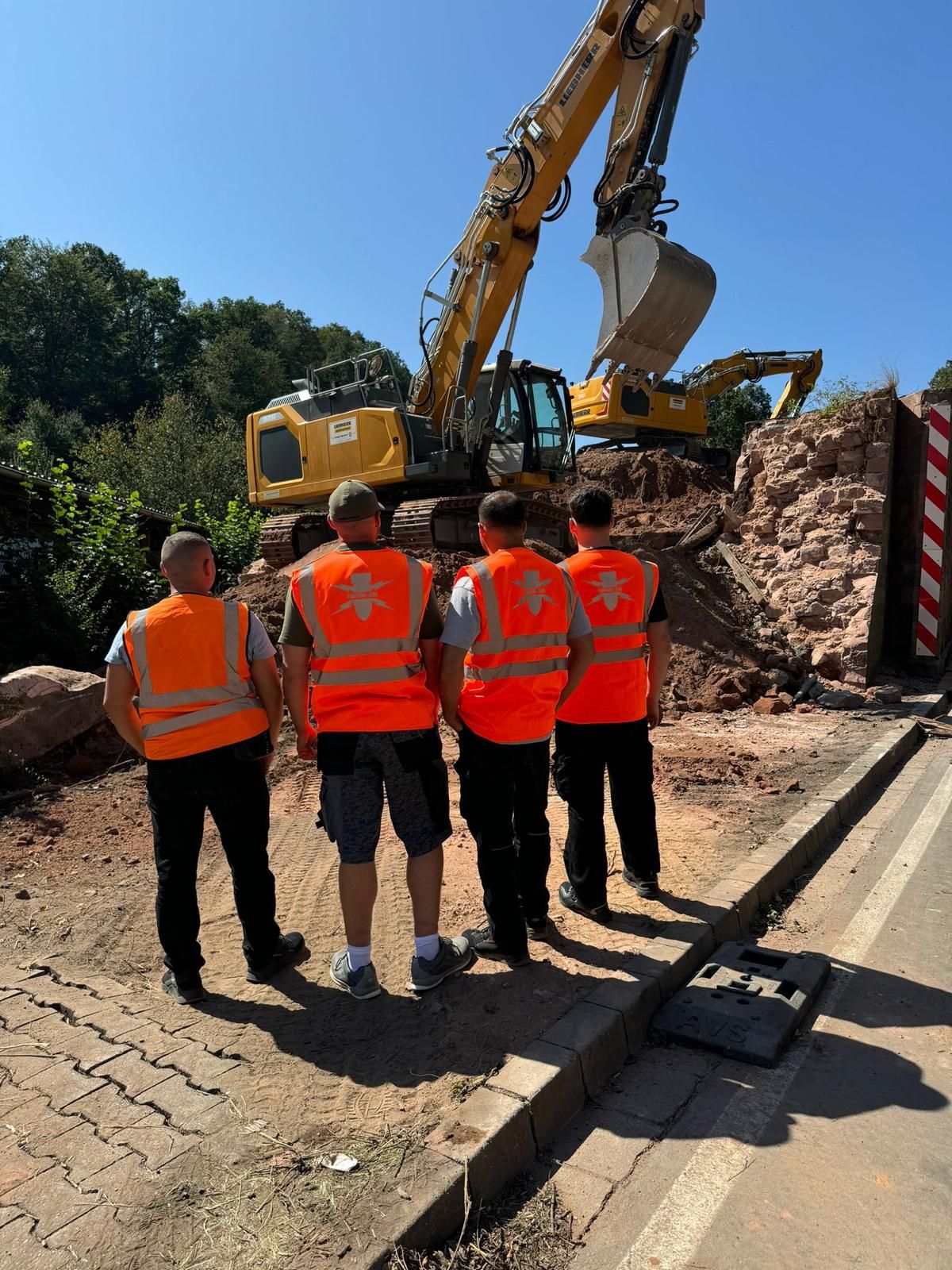 Vier Personen in orangefarbenen Warnwesten vor zwei Baggern auf einer Baustelle.
